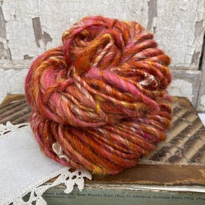 May include: A thick, hand-spun yarn skein featuring a variegated blend of vibrant orange, deep red, and bright pink fibers. The yarn has a textured, multi-ply appearance with subtle variations in color intensity throughout the twisted bundle.