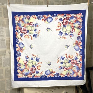 May include: A square white fabric cloth featuring a wide dark blue border and a central pattern of colorful fruit clusters. The design includes illustrations of blue, red, and yellow plums, grapes, and leaves arranged along the inner border.