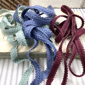 May include: Close-up of three different colors of decorative trim. The trim is in shades of blue, green, and burgundy. The trim is made of a woven material with a scalloped edge. The trim is laying on top of an old book.