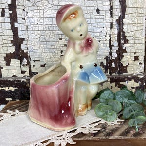 May include: A vintage ceramic figurine of a child in a hat and blue outfit, holding a maroon shoe-shaped planter. The figurine has a pink bow and is set against a distressed white and brown background. Decorative greenery is also present.