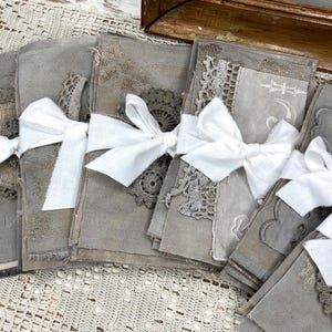 May include: A collection of handmade journals tied with white linen bows. The journals are a muted gray color, with decorative lace and fabric accents. The overall aesthetic is vintage and rustic.