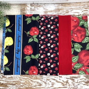 May include: A collection of fabric swatches featuring fruit and text designs. The fabrics include patterns of red apples, yellow pears, and solid red. One swatch has text that reads "Cortland - Northern Spy - Jonathan - McIntosh - Red Delicious".