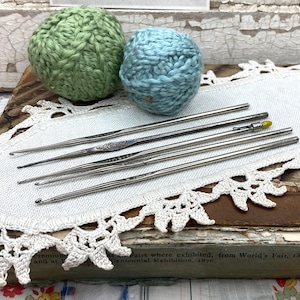 May include: A collection of crochet hooks and two yarn balls. The yarn balls are light green and blue. The crochet hooks are silver and are arranged on a white lace doily. The doily is on top of a vintage book.