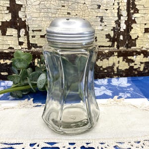 May include: A clear, vintage glass salt shaker with a silver metal lid. The shaker has a faceted, hourglass-like shape. The background includes a distressed wooden surface and a blue and white patterned cloth.