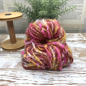 May include: A skein of thick, textured yarn featuring a variegated blend of deep magenta, mustard yellow, and white fibers with a fuzzy, irregular appearance. Beside the yarn sits a light brown wooden spool with a circular base and top.