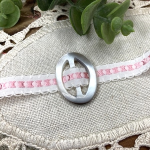May include: Oval-shaped, iridescent button with a cut-out design, resting on a white ribbon with pink embroidered details. The button is centered on a textured, beige fabric, with green foliage in the background. A delicate craft item.