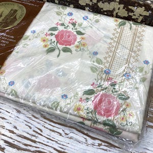 May include: A twin size bed sheet in original plastic packaging featuring a cream background with a floral pattern of pink roses, yellow blossoms, and blue flowers, accented by green leaves and a vertical tan dotted border stripe design.