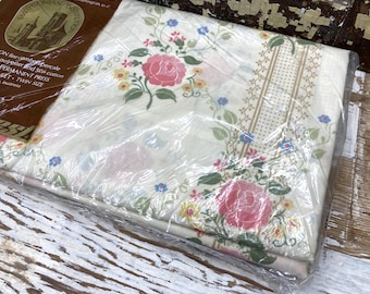 May include: A twin size bed sheet in original plastic packaging featuring a cream background with a floral pattern of pink roses, yellow blossoms, and blue flowers, accented by green leaves and a vertical tan dotted border stripe design.