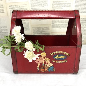 May include: A vintage red metal toolbox with a handle, featuring a colorful illustration of children and the text "SHU-SHINE BANK Amsco For Kiddies." White flowers and green stems are placed inside the toolbox.