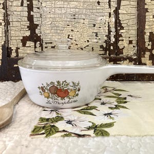 May include: A white ceramic saucepan with a clear glass lid and a long handle. The side of the pan features a colorful illustration of various vegetables and fruits, including a tomato, with the cursive text La Marjolaine printed below.