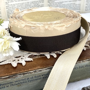 May include: A roll of tan and brown ribbon. The ribbon is wrapped around a cardboard core with a dark brown band. A length of the ribbon extends outward. The image includes a white flower and text that reads "First Premiums awarded at all Fairs where".