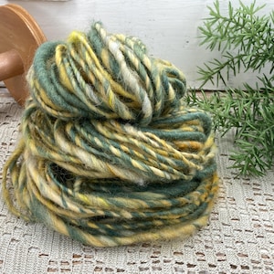 May include: A ball of hand-spun yarn in shades of green, yellow, and cream. The yarn is thick and textured, with a rustic appearance. A wooden spindle is partially visible in the background. The yarn is likely used for knitting or crocheting.