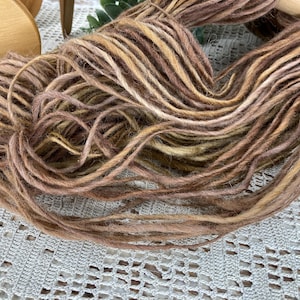 May include: Close-up of a skein of hand-dyed yarn in shades of brown, tan, and cream. The yarn is loosely wound, showing the texture and color variations. The yarn is likely for knitting or crochet projects.
