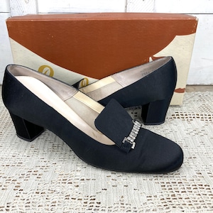 May include: A pair of black satin heeled shoes with a square toe and a decorative rhinestone buckle. The shoes have a block heel and are displayed with their original box, which has a brown and cream design.