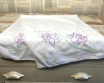 May include: A white rectangular table runner featuring embroidered purple orchid flowers and green swirling stems. The fabric edges are finished with a delicate white lace trim. The embroidery is centered on the runner, showcasing detailed floral patterns.