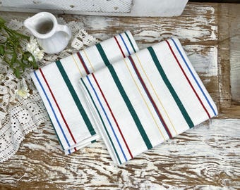May include: Two white kitchen towels with vertical stripes in red, green, blue, and yellow. The towels are folded and placed on a rustic wooden surface. A small white pitcher is visible.