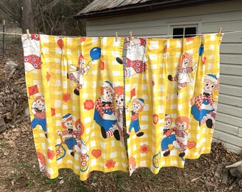 May include: A pair of yellow and white gingham curtains with a repeating pattern of Raggedy Ann and Andy dolls. The curtains have a gathered top and are hung on a clothesline.