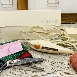 May include: Vintage Singer sewing machine accessories, including a cream-colored case, iron, and scissors. A red pincushion with a green leaf and a variety of fabric scraps are also present. The Singer logo is visible on the case and iron.