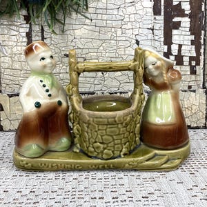 May include: A vintage ceramic wishing well figurine featuring a boy and girl, with the words "Wishing Well" on the base. The well is olive green, and the figures are dressed in brown, white, and green. A charming decorative piece.
