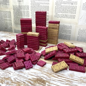 May include: A collection of rectangular wooden dominoes in shades of deep red and natural tan. The set includes several vertical stacks of dominoes and a large pile of individual pieces scattered across the surface, each featuring indented pips.