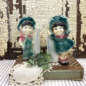 May include: Two vintage ceramic figurines of children, one a boy in overalls and the other a girl in a dress, both wearing hats. The figurines are painted in shades of green, red, and white. They are displayed on a wooden base with a lace doily.