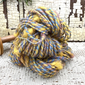 May include: A large ball of colorful yarn in shades of yellow, blue, and purple. The yarn is thick and textured, with a slightly fuzzy appearance. A wooden spindle is visible in the background, suggesting the yarn is for crafting.