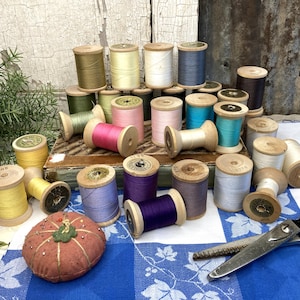 May include: A collection of vintage wooden spools of thread in various colors, including yellow, pink, blue, and purple. A round, orange pincushion and a pair of scissors are also present. The items are arranged on a blue and white checkered cloth.