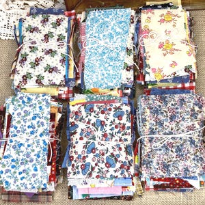 May include: A collection of fabric bundles tied with white string. The bundles feature various floral patterns in shades of blue, red, yellow, and white. The fabrics are arranged on a neutral-toned surface, creating a visually appealing display.