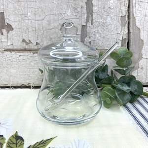 May include: A clear glass jam jar with a matching lid and a glass spoon. The jar features a delicate etched floral pattern around its body. The lid has a small knob handle, and the jar includes a side notch to accommodate the included glass spoon.