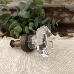 May include: A clear, faceted glass cabinet knob with a multi-sided geometric shape. The knob features a dark, aged bronze-toned metal base and a short, cylindrical metal stem extending from the back for mounting purposes.