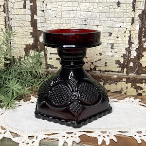 May include: A dark red glass candle holder with a square base and a round top. The candle holder has a textured, patterned design on the base and stem. The top is a deep red color, and the holder is sitting on a white doily.