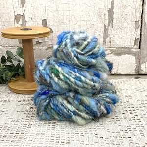May include: A thick, textured skein of yarn featuring a variegated blend of light blue, dark blue, grey, and white fibers. The yarn has a chunky, uneven ply with visible fuzzy strands and is positioned next to a wooden spool with a round base.