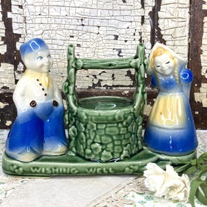 May include: A vintage ceramic wishing well figurine with two figures. The well is green, and the figures are dressed in blue and white. The base reads "Wishing Well". The figurine is set against a distressed, white-painted wooden background.