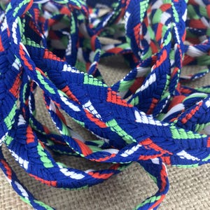 May include: A close-up of a blue, red, white, and green braided elastic trim. The trim has a geometric pattern and is perfect for adding a pop of color to any project.