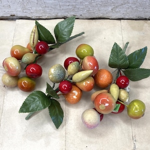 May include: Three decorative fruit clusters featuring artificial pears, cherries, and oranges with green fabric leaves. The fruits have a glossy, multi-colored finish in shades of red, orange, yellow, and green, attached to thin stems.