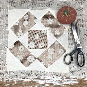 May include: A handmade quilt block with a floral pattern in brown and white, surrounded by white triangles. A red tomato-shaped pin cushion and a pair of scissors are next to the quilt block. The items are on a lace doily.