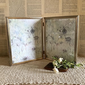 May include: A double-hinged gold metal picture frame containing two identical prints of line-drawn flowers with soft watercolor accents in shades of green, blue, and purple. The frame is positioned upright and features a thin, metallic border.