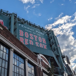 Könnte beinhalten: Ein grünes und weißes Schild mit dem Text "Fenway Park, Home of the Boston Red Sox" vor einem blauen Himmel mit weißen Wolken.