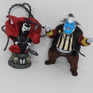 May include: Two action figures from the Spawn comic book series. One is a bust of Spawn with a red cape and skull base. The other is a clown-like character with a blue face, black jacket, and brown pants. Both are connected by a chain.