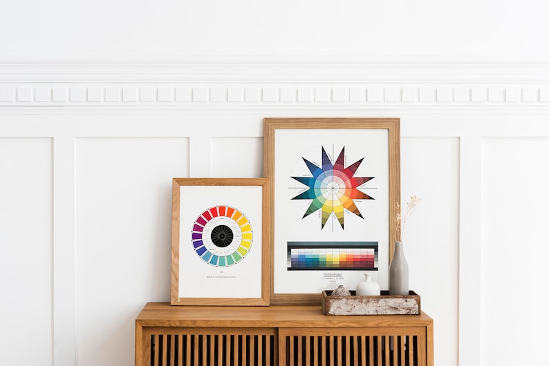 May include: Two framed color wheel charts hang on a white wall above a wooden cabinet. The chart on the left is a circular color wheel with 12 colors. The chart on the right is a star-shaped color wheel with 12 colors and a color bar below it.
