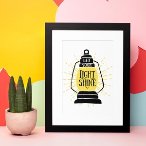 May include: A black framed print with a white background featuring a yellow lantern with the text "Let your light shine" in black lettering.