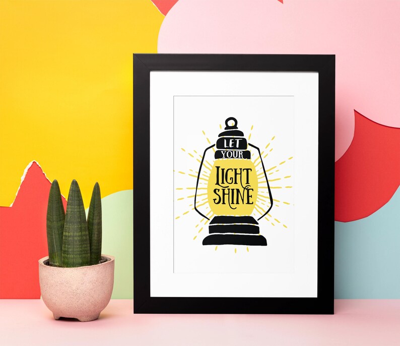 May include: A black framed print with a white background featuring a yellow lantern with the text "Let your light shine" in black lettering.