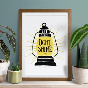 May include: A framed print with a white background and a black silhouette of a lantern with the text "Let your light shine" in yellow lettering.