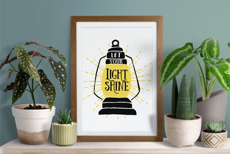 May include: A framed print with a white background and a black silhouette of a lantern with the text "Let your light shine" in yellow lettering.