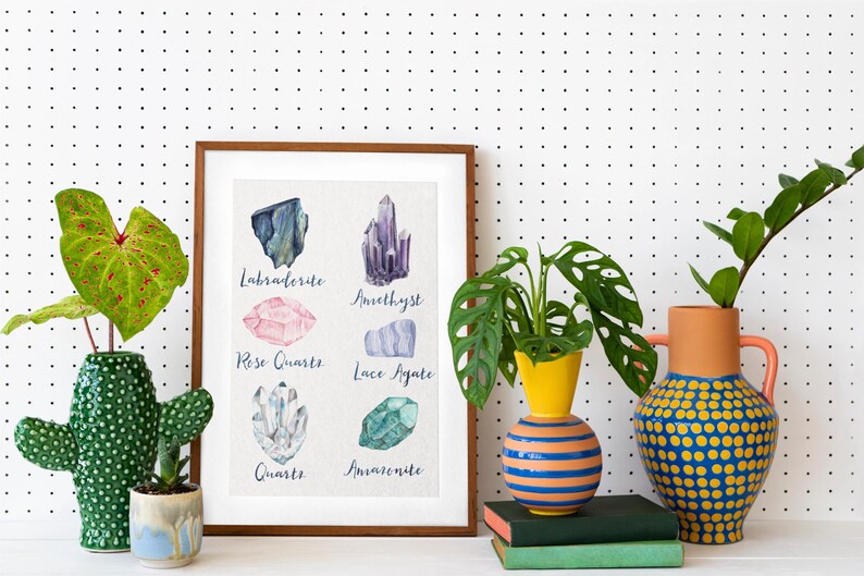 May include: A framed print featuring watercolor illustrations of various crystals, including Labradorite, Amethyst, Rose Quartz, and Quartz. The print is displayed with decorative vases and potted plants against a white backdrop with black polka dots.