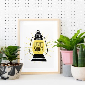 May include: A framed print with a black and yellow illustration of a lantern with the text "Let your light shine". The print is displayed on a white pegboard wall with several potted plants in front of it.