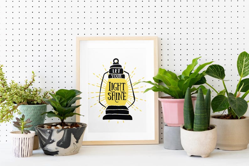 May include: A framed print with a black and yellow illustration of a lantern with the text "Let your light shine". The print is displayed on a white pegboard wall with several potted plants in front of it.