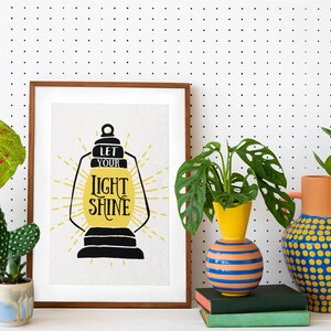 May include: A framed print with the text "Let your light shine" in a yellow lantern design. The print is displayed on a white pegboard wall with three potted plants in front of it.