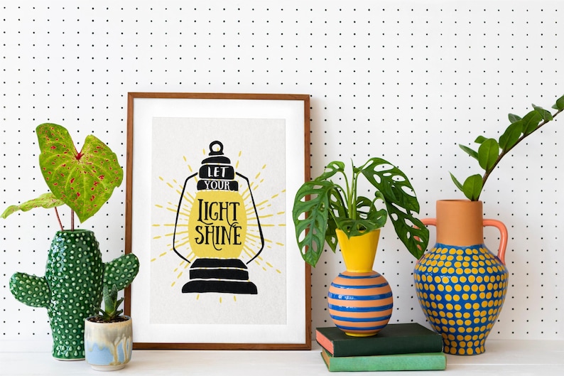 May include: A framed print with the text "Let your light shine" in a yellow lantern design. The print is displayed on a white pegboard wall with three potted plants in front of it.