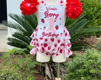 Könnte beinhalten: Ein rosa-weißer Strampler mit Erdbeermotiv und dem Text "Ty'Lasia's Berry 1st Birthday". Der Strampler hat einen gerüschten Rock und ist mit roten Rosetten und einer rosa Schleife verziert. Das Outfit wird auf einer Schaufensterpuppe präsentiert.